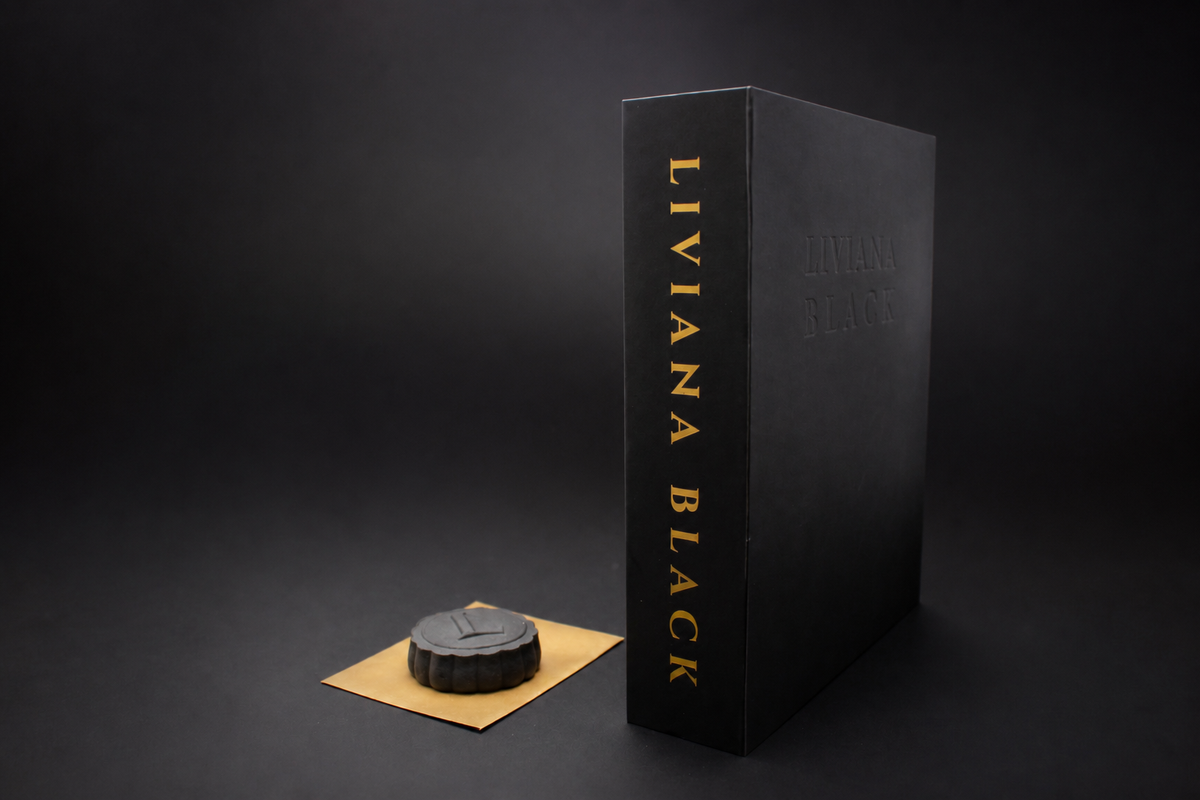 A black rectangular book box with gold vertical text on spine reading 'LIVIANA BLACK' stands on a dark studio background, next to a small round black mooncake with an embossed L resting on a gold rectangular envelope.