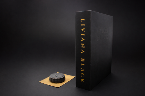 A black rectangular book box with gold vertical text on spine reading 'LIVIANA BLACK' stands on a dark studio background, next to a small round black mooncake with an embossed L resting on a gold rectangular envelope.