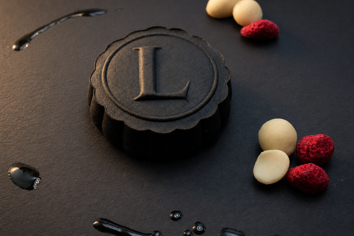 A round black sesame mooncake with a scalloped edge and a large embossed letter 'L' in the center, styled on a dark textured surface with macadamia nuts and raspberries decorated around it and black sesame paste drizzled as well