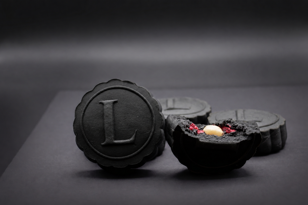 four black round mooncakes with the front mooncake featuring an embossed L inside an embossed circle with two full mooncakes in background and a mooncake broken open to reveal red raspberry and macadamia nut halves inside with black sesame filling