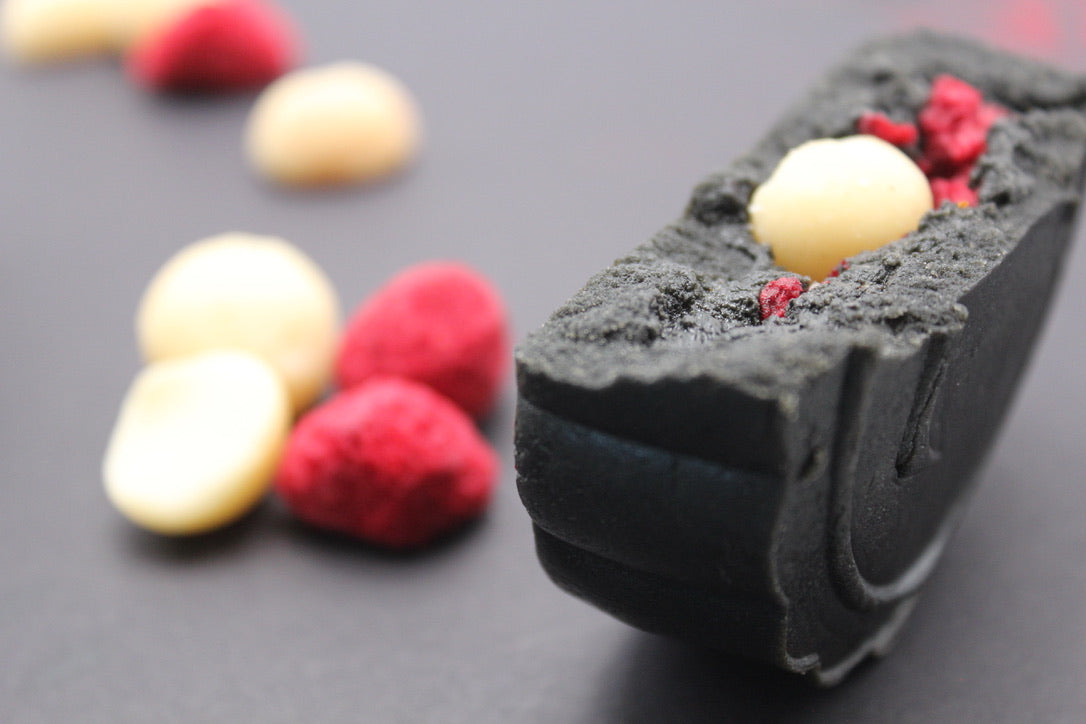 black mooncake half showing raspberry and macadamia pieces with black sesame filling; red whole raspberries and macadamia halves are in the background