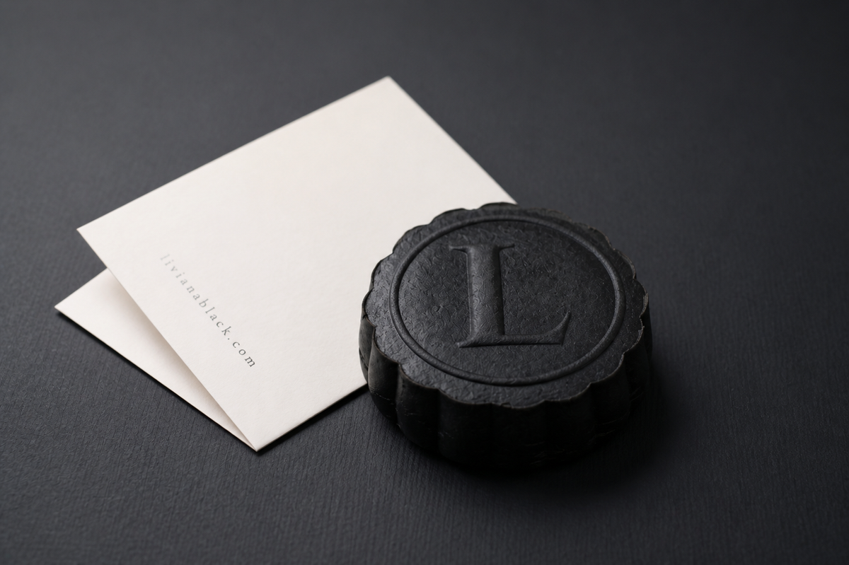 A round black sesame mooncake with a scalloped edge and an embossed letter 'L' on top, shown next to a cream colored folded card on a dark textured background.