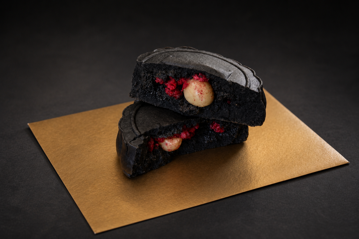 Two halves of a black sesame mooncake with red raspberry and macadamia and black sesame filling, stacked on a rectangular gold envelope against a dark background.