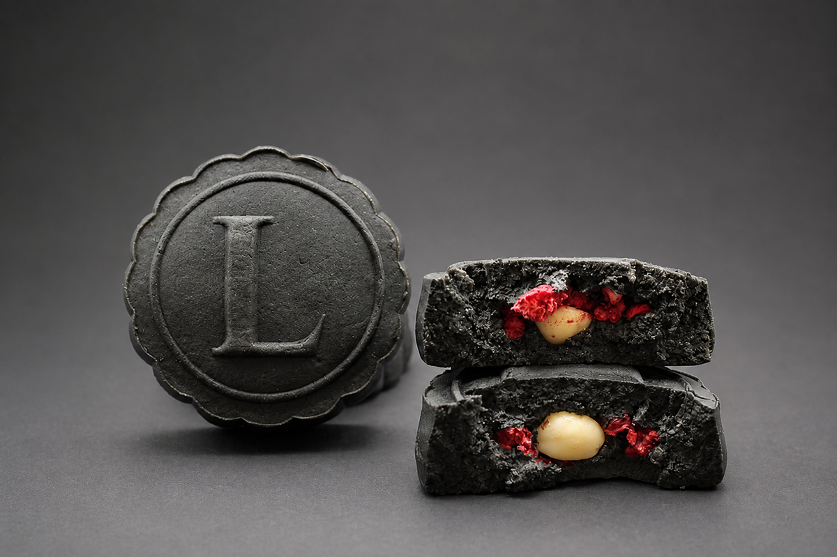 A black mooncake with an embossed letter 'L' stands next to two broken open mooncake halves showing a textured interior filled with red raspberries, macadamia nut halves, and black sesame paste filling, set against a dark gray background.