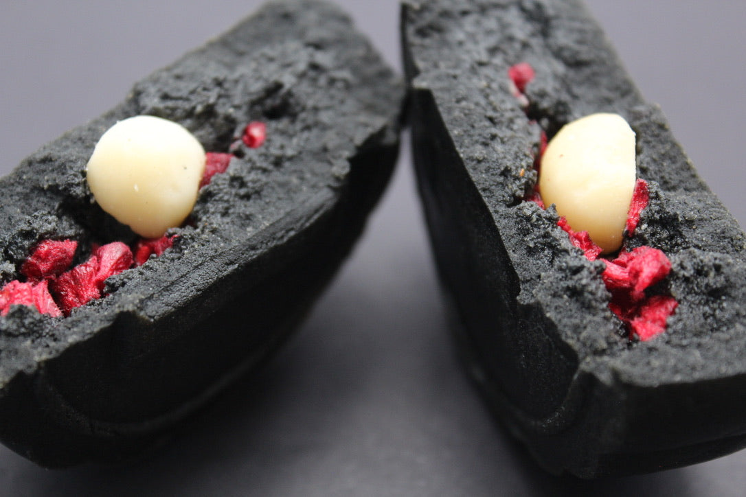 A close-up of a two broken open halves of a black sesame mooncake, showing creamy macadamia nut halves, black sesame paste filling, and bright red raspberry pieces